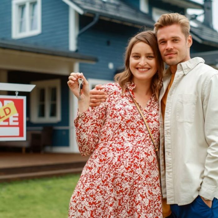 Portrait of a Beautiful Young Couple in Love Standing in Front Their New Home. Successful Homeowners Looking at Camera and Smile. Female in a Dress Expecting a Baby. Real Estate Housing Market Concept